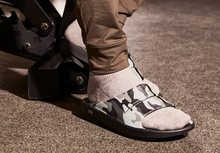 Load image into Gallery viewer, A close-up of a person's foot wearing a gray knit sock and a black slide sandal with a gray and white camouflage strap, standing on a textured gray floor next to a black mechanical device.