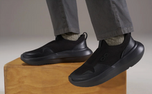 Load image into Gallery viewer, A pair of black slip-on athletic sneakers worn with gray pants and socks, with one shoe resting on a wooden block and the other suspended in midair against a neutral studio background.
