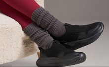 Load image into Gallery viewer, A person wearing black slip-on mesh sneakers with thick black soles, paired with gray ribbed leg warmers and red leggings, seated on a cream textured chair.
